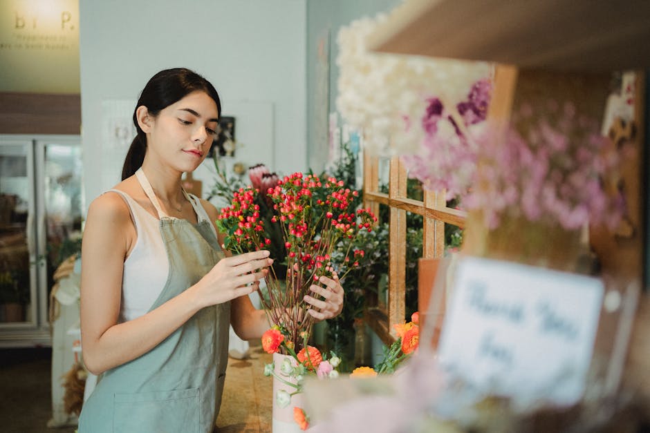A young woman in a light-colored apron is selecting a bouquet of small, bright pink and red hypericum berries with green stems and leaves, standing behind a floral counter inside a flower shop. The ar