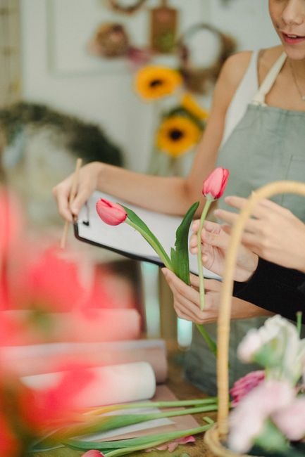A person holding a smartphone and preparing a floral arrangement featuring fresh pink tulips with green stems and leaves, set on a worktable decorated with wrapping paper and floral tools. In the back