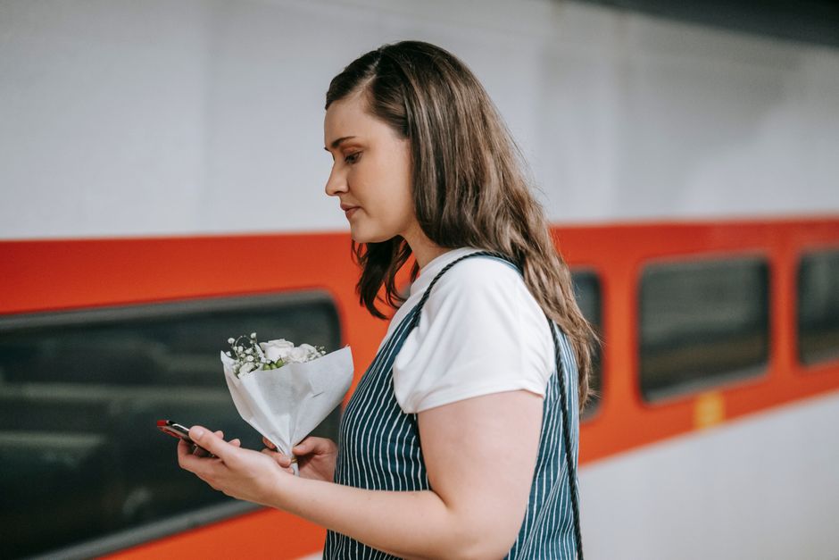 A woman with shoulder-length brown hair, wearing a white t-shirt and a blue and white striped apron, stands on a train platform holding a bouquet of fresh white flowers with green foliage wrapped in w