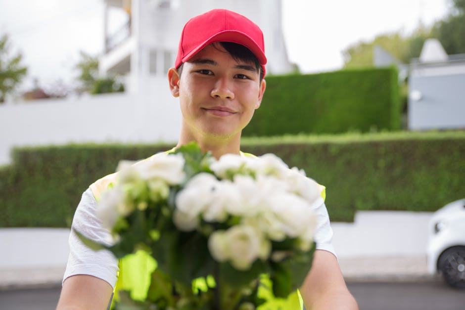 A delivery person wearing a red cap and a high-visibility vest extends a bouquet of fresh, white roses and green foliage towards the camera, standing outdoors on a residential street. Behind them, the