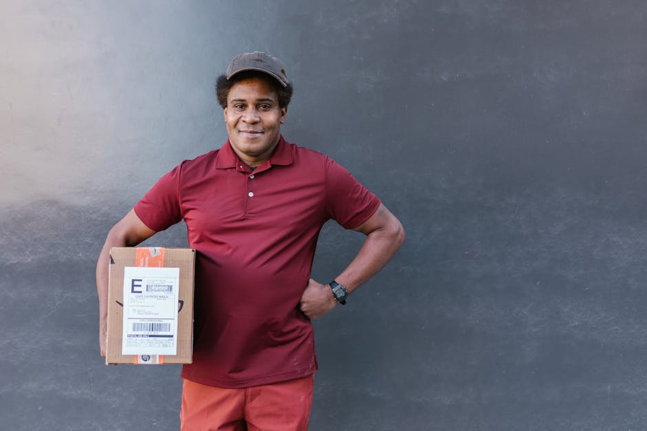 A smiling delivery person in a burgundy polo shirt, red pants, and a dark cap stands outdoors in front of a plain dark grey wall, holding a small cardboard parcel with a shipping label. The individual