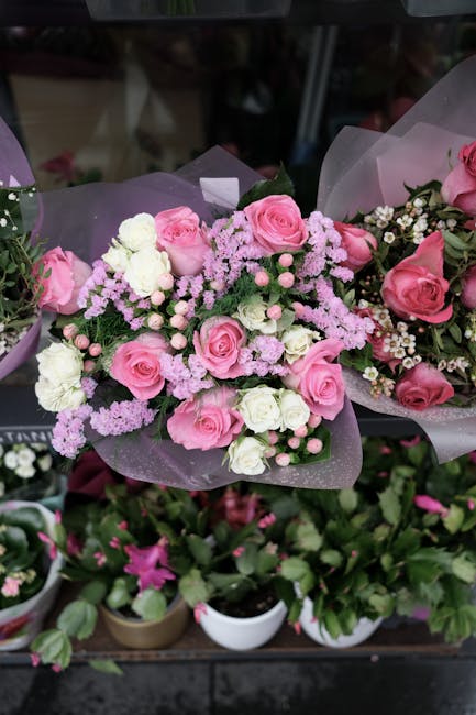 A floral arrangement featuring a bouquet of fresh pink roses, white spray roses, and lavender-pink astilbe, all meticulously arranged with green foliage and wrapped in soft purple tissue paper. The bo