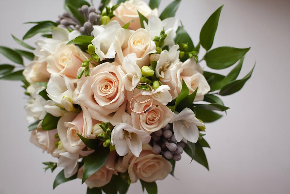 A close-up view of a fresh floral bouquet arranged with pale pink roses, white freesias, and small clusters of grey berries, complemented by glossy green leaves. The bouquet is elegantly bound togethe