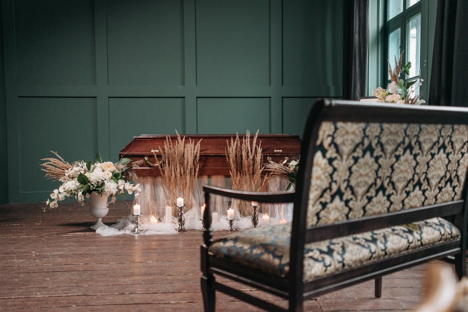 A wooden funeral casket placed on a floor with dark driftwood planks, decorated with dry pampas grass and pale pink and white floral arrangements in white vases. Surrounding the casket are small silve